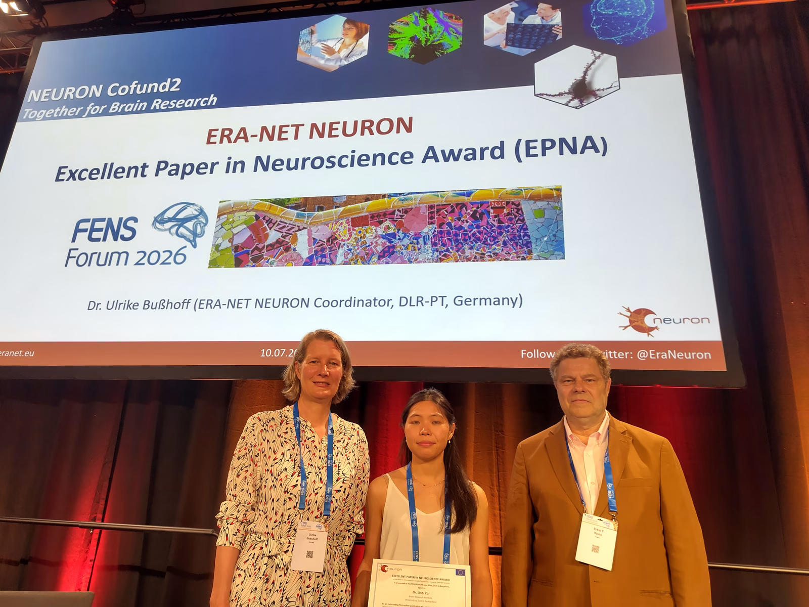 Three people wearing conference badges stand on a stage in front of a large screen displaying the ERA-NET NEURON Excellent Paper in Neuroscience Award (EPNA) presentation slide