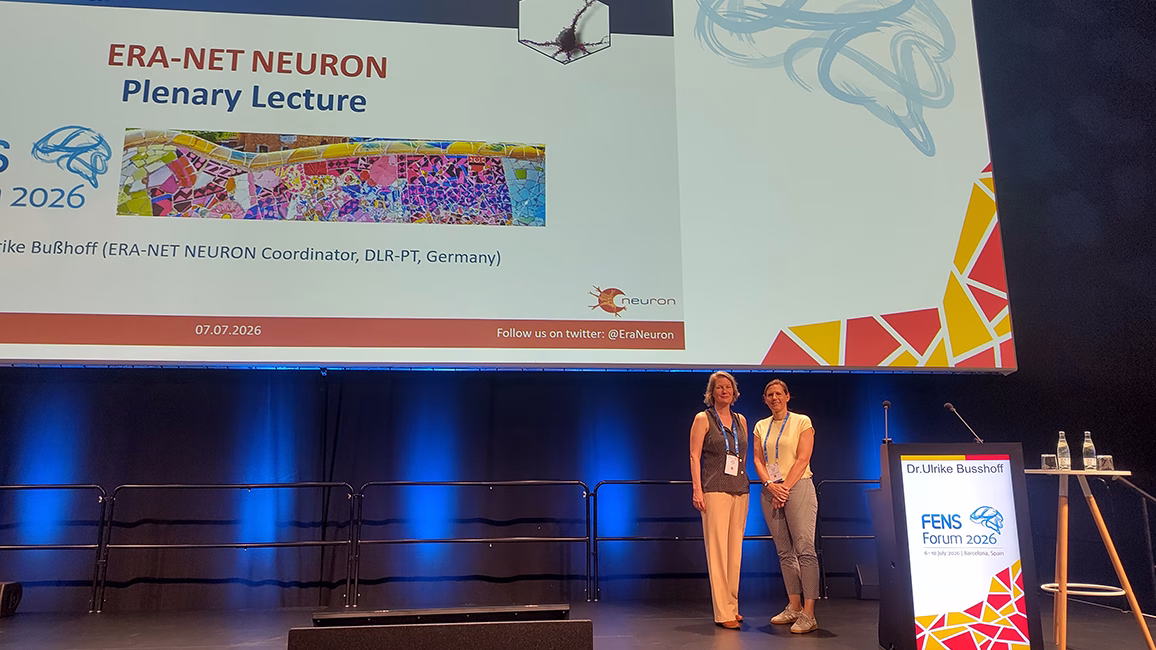 Two women standing on a stage next to a lectern with a presentation slide titled 'ERA-NET NEURON Plenary Lecture' projected behind them