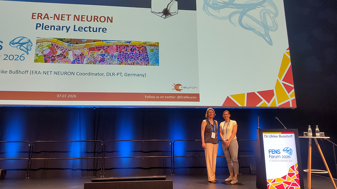Two women standing on a stage next to a lectern with a presentation slide titled 'ERA-NET NEURON Plenary Lecture' projected behind them