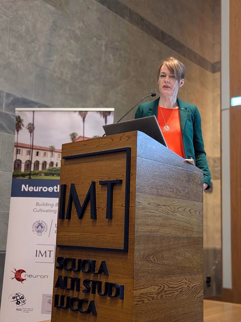 Person standing behind a wooden podium with 'IMT Scuola Alti Studi Lucca' on the front, speaking in a room with a stone wall and a banner showing a building and palm trees