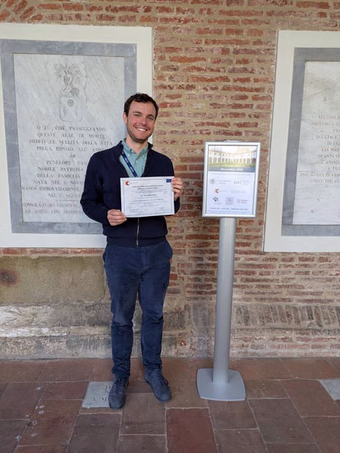 Person standing indoors against a brick wall holding a certificate next to a sign on a stand