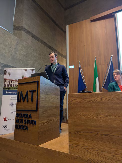 Person standing behind a wooden podium with 'IMT' and 'SCUOLA DI STUDI ITALIANI' on it in a conference room with flags and a seated person at a wooden desk